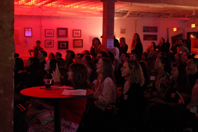A diverse group of people sits closely together in a dimly lit room with red lighting, smiling and watching something off-frame.